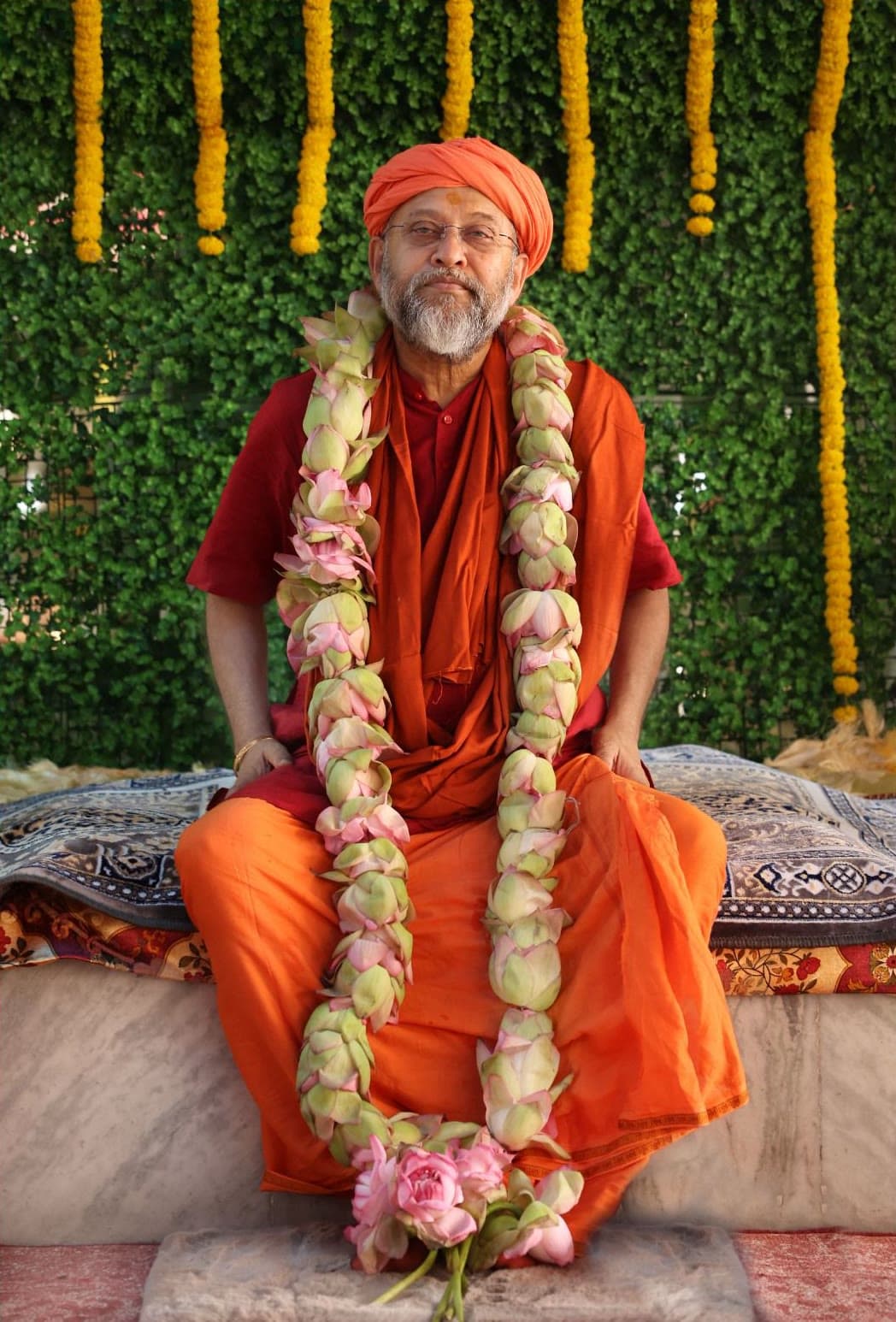 Swami Debananda Maharaj seated at a recent darshan, wearing a saffron turban and a lotus garland