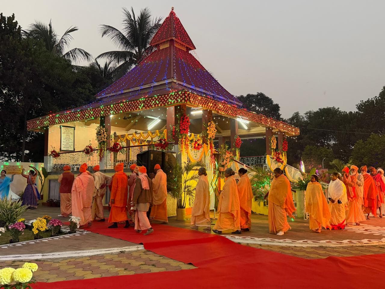 An evening procession of monks and devotees in saffron along a red carpet, past a temple pavilion strung with coloured lights