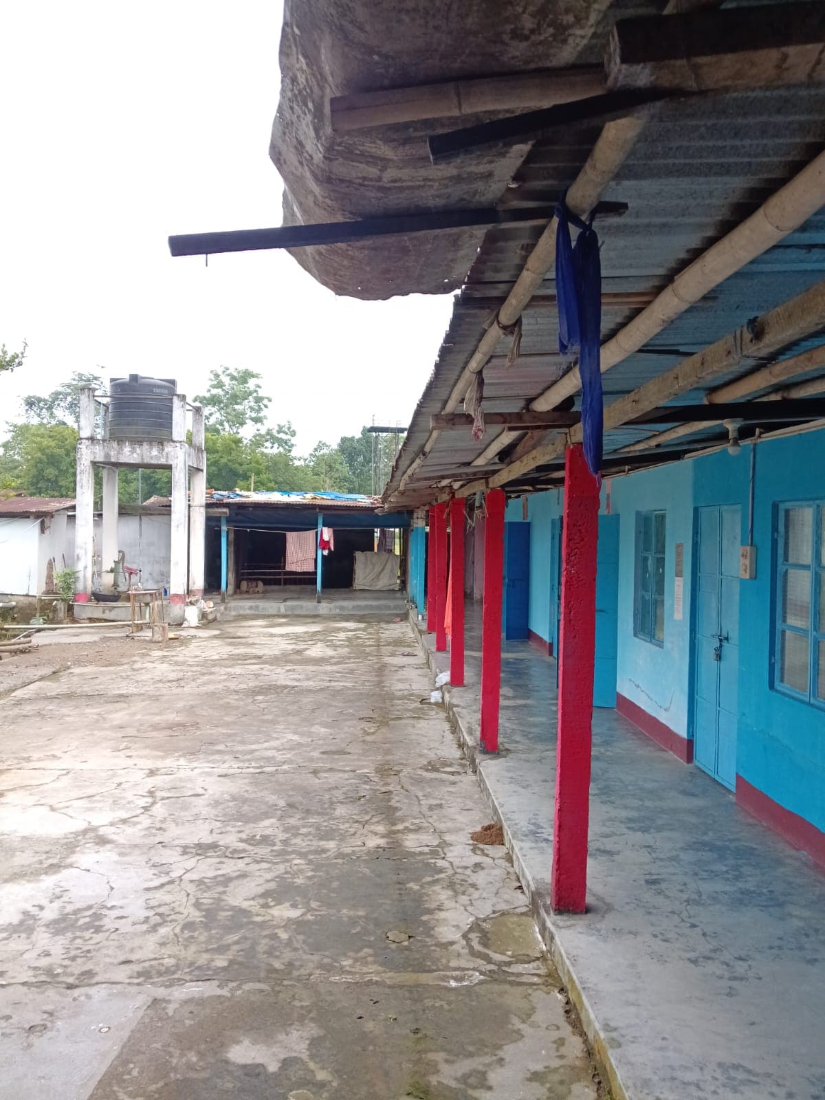 A long veranda with red pillars and blue walls running along a row of rooms, with a water tank in the distance
