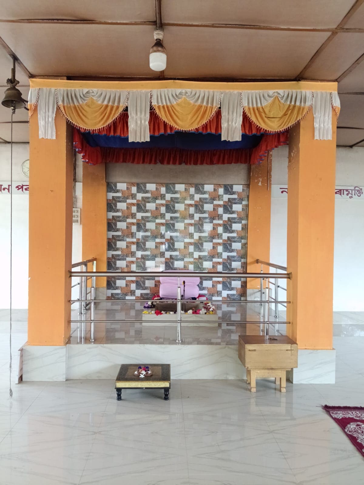 An altar framed in orange pillars and draped curtains, with a low wooden stool on the tiled floor in front