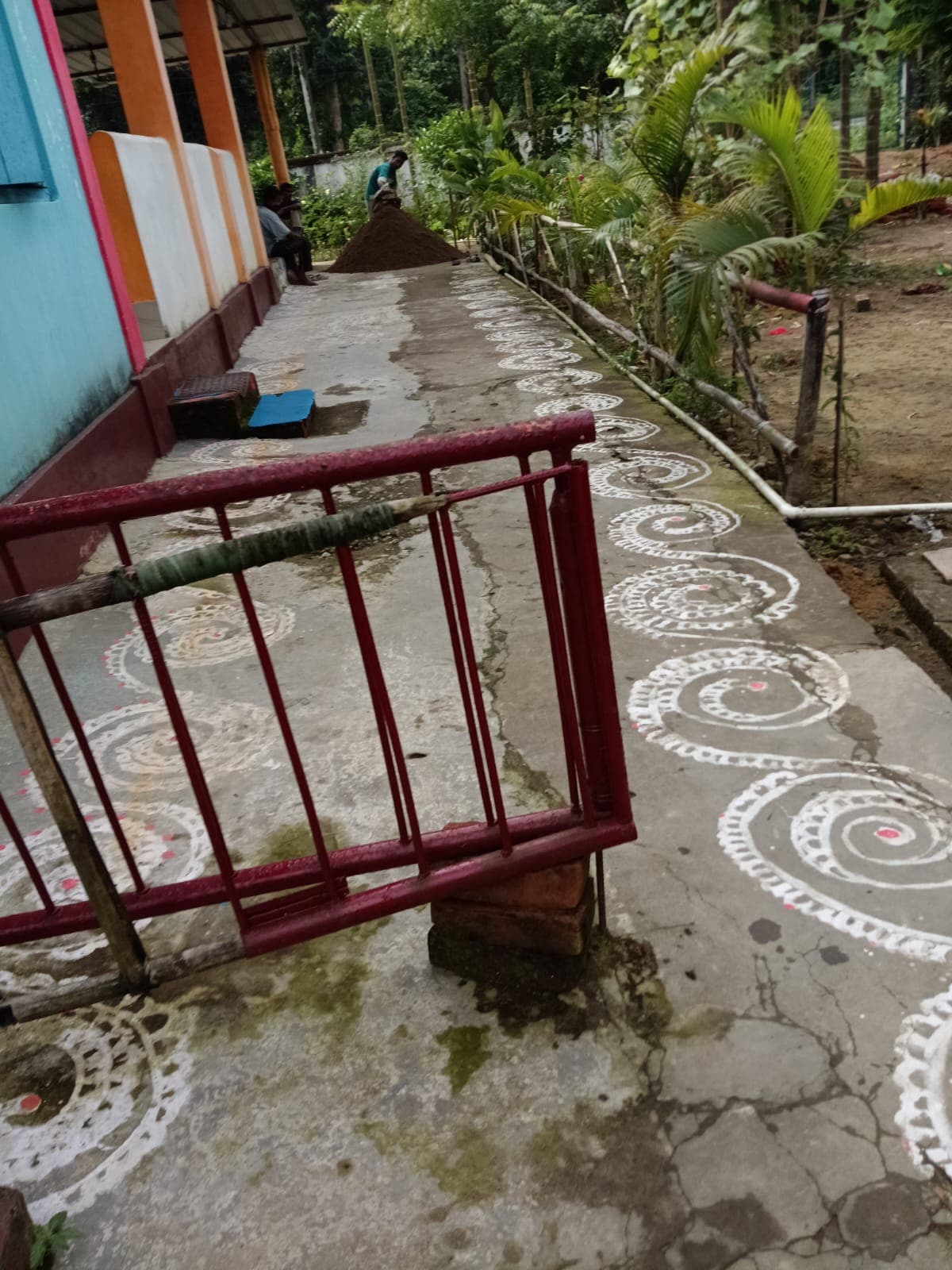 A whitewashed path decorated with swirling alpana patterns beside a garden, with a red railing in the foreground