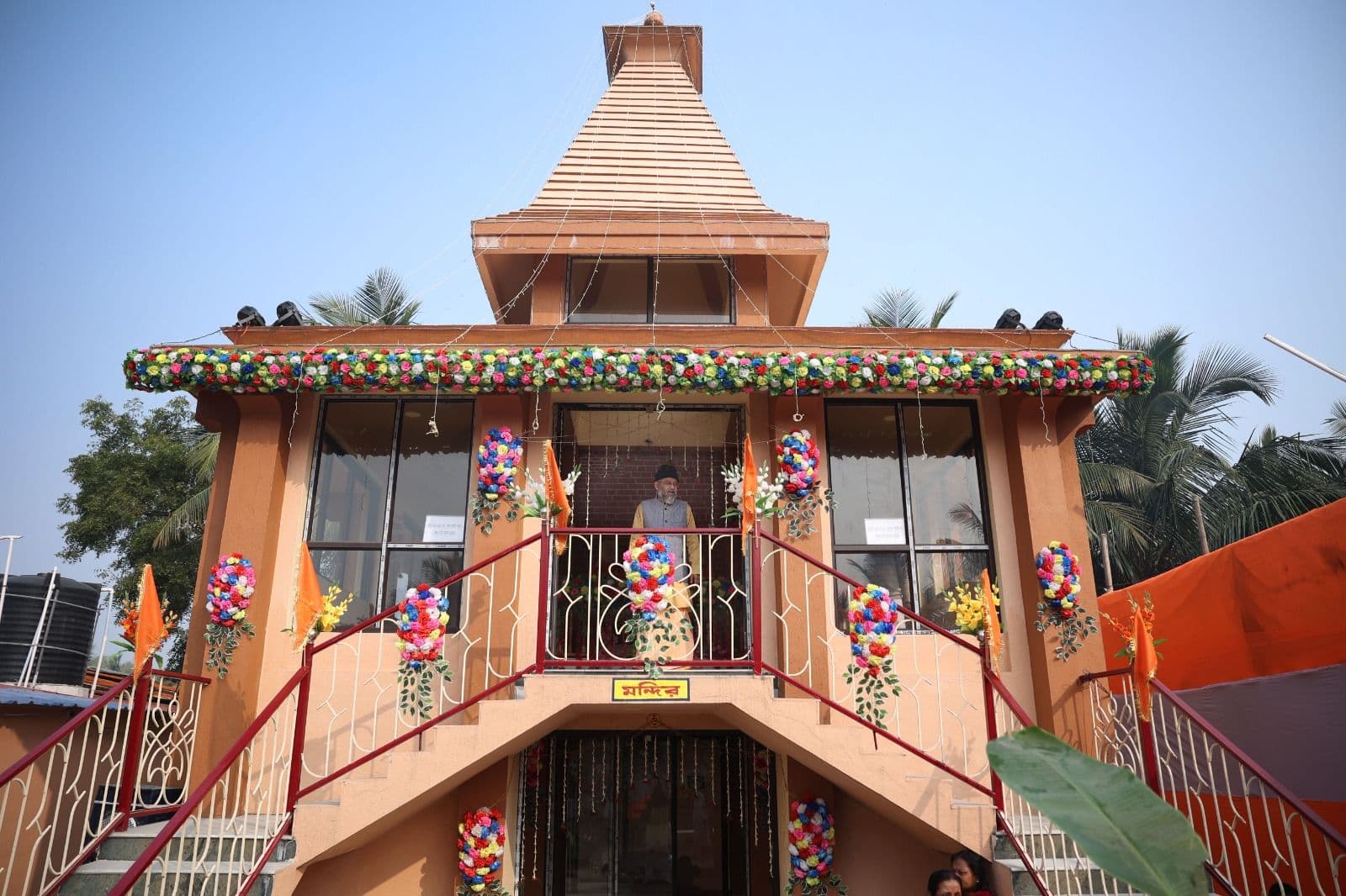 A tiered orange temple building decorated with flowers along its balcony, with twin staircases rising to the entrance