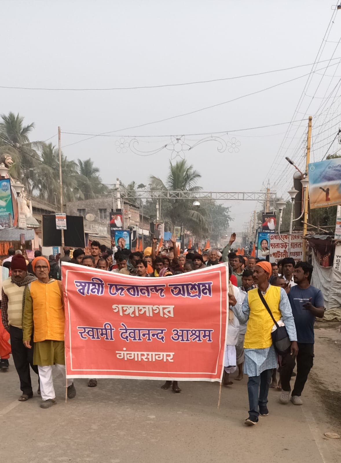 A procession of people in saffron and yellow walking down a road behind a red banner lettered in Bengali