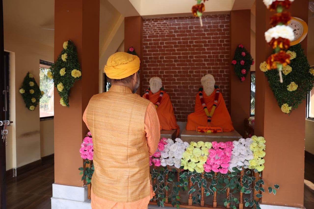 A man in a saffron turban standing before a shrine with two seated statues, framed by flower garlands