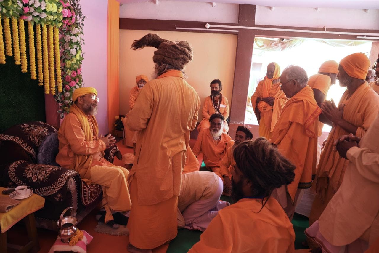 A gathering of men in saffron robes and turbans, some seated and some standing, inside a room hung with garlands