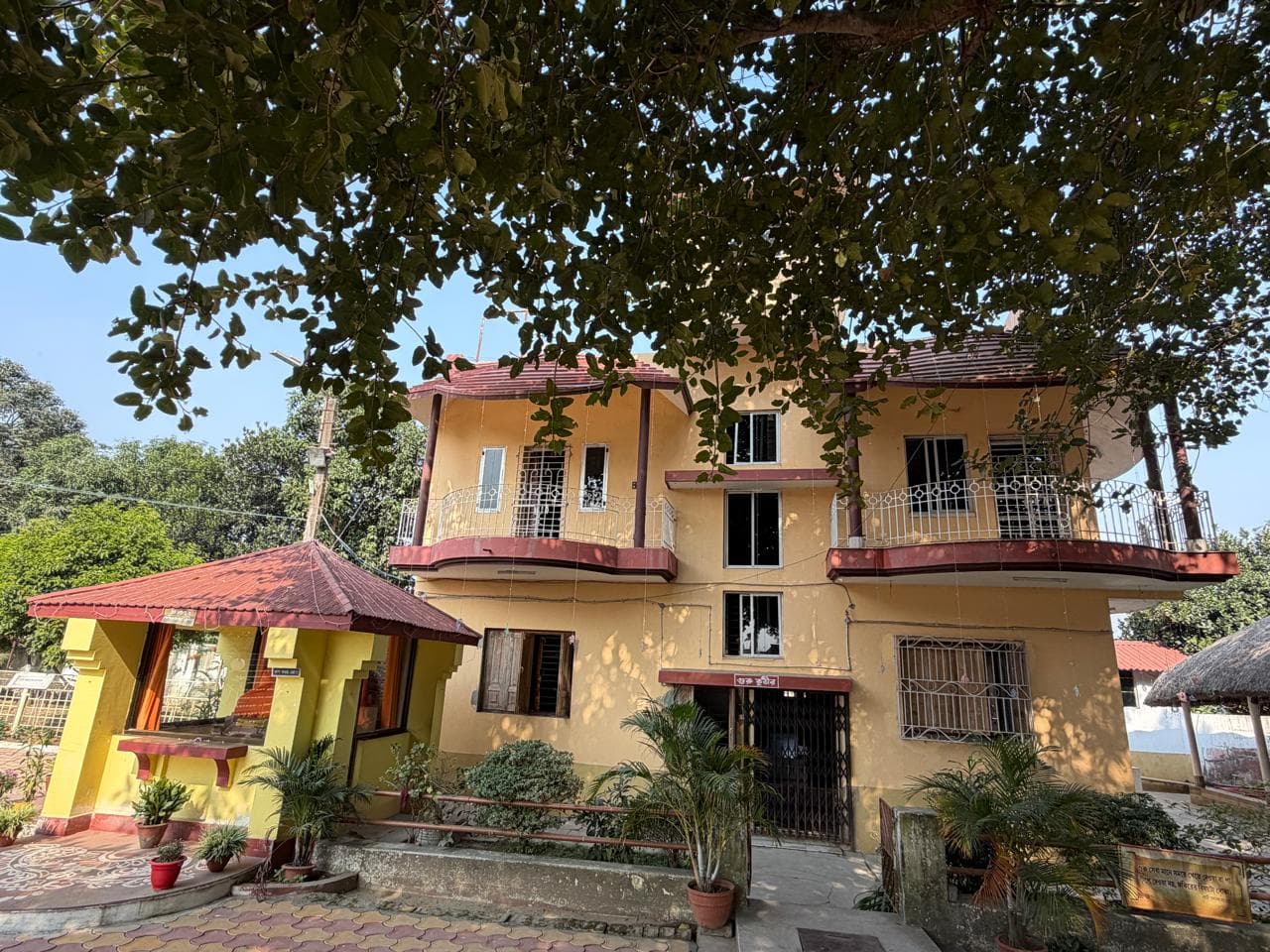 A yellow two-storey building with red balconies and a small red-roofed shrine in front, seen beneath a tree