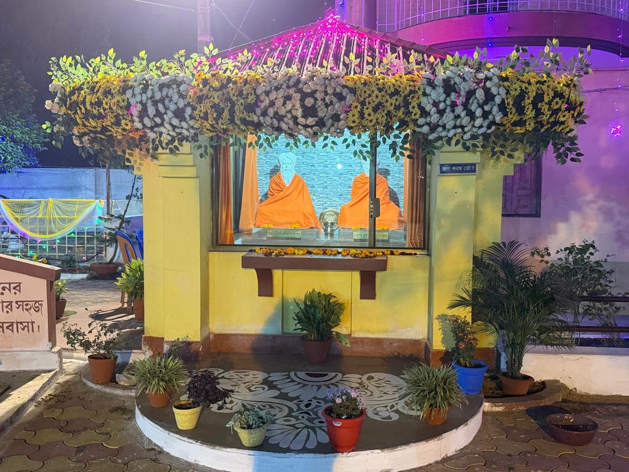 A small yellow shrine with a window showing two seated figures in saffron, crowned with flowers and lit purple at night