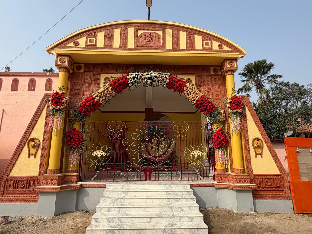 An ornate arched shrine painted red and yellow, its entrance garlanded with red and white flowers above white marble steps