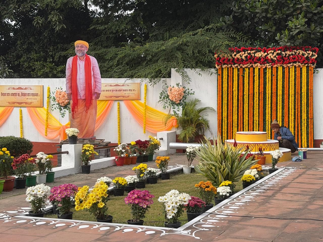 A tall standing figure in saffron beside a flower garden and a backdrop hung with marigold garlands