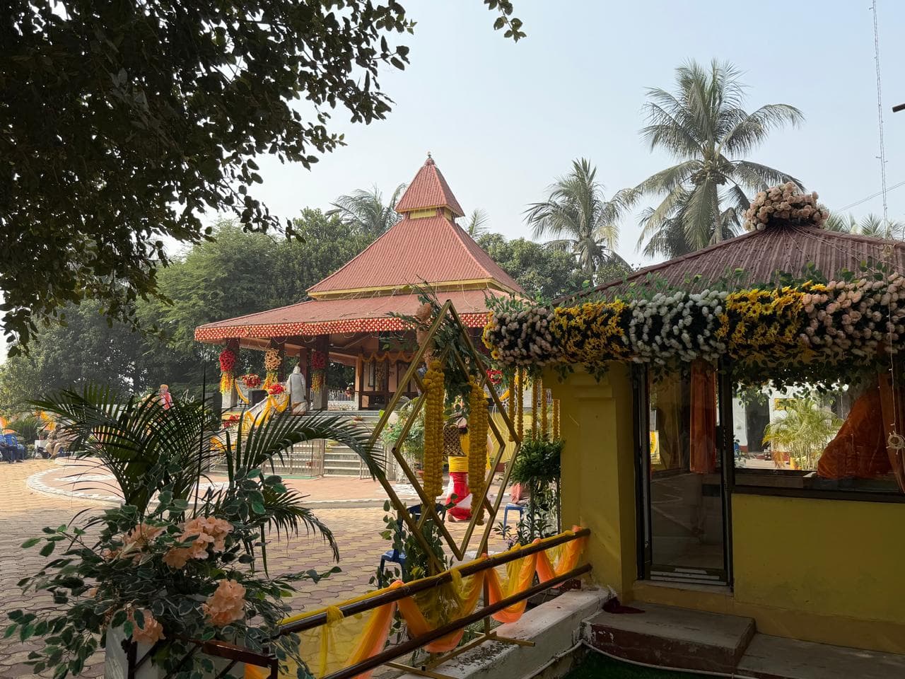 A red-roofed open temple pavilion across a courtyard, seen past a small shrine decorated with flowers