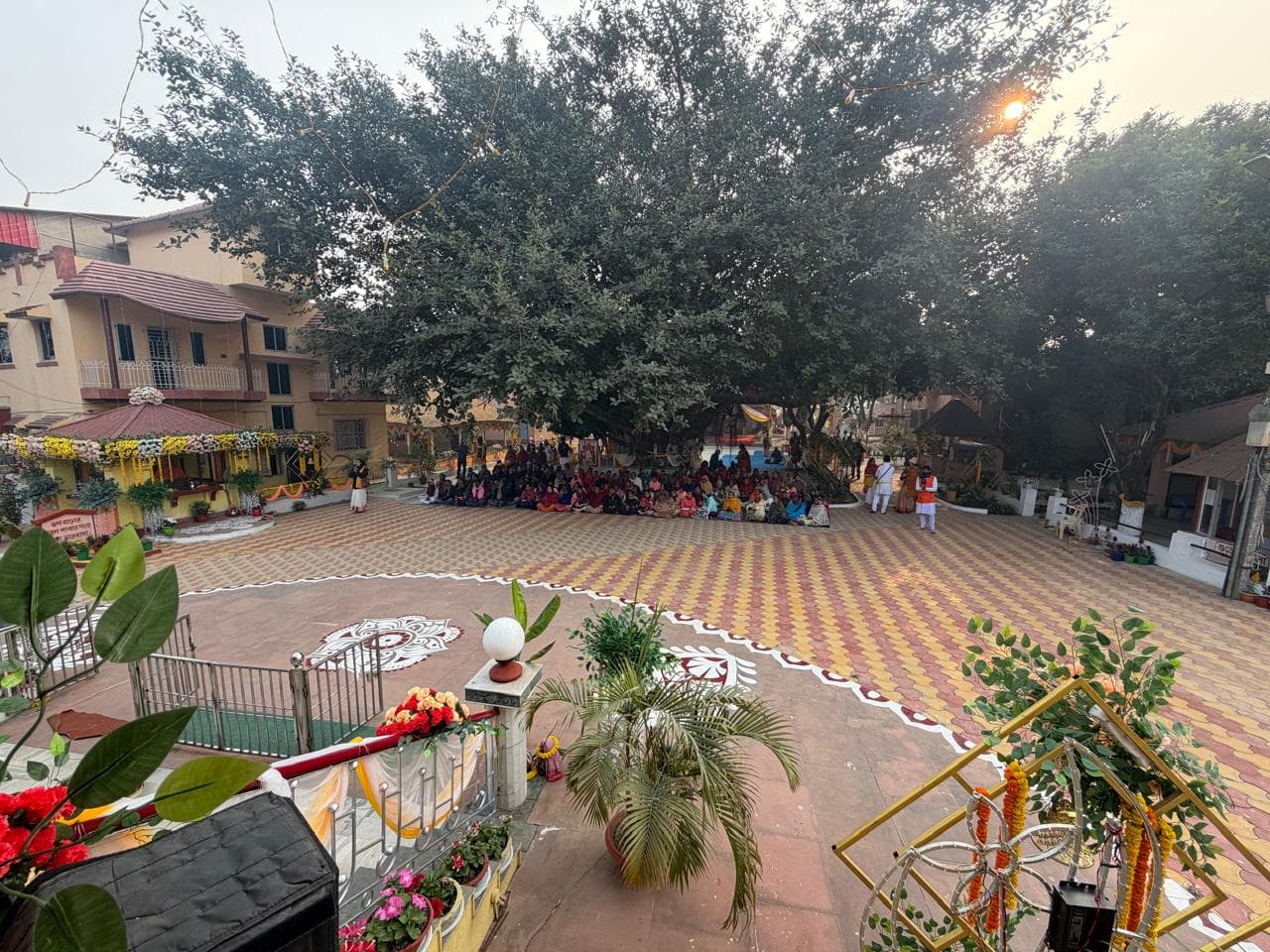 A wide paved courtyard with alpana patterns on the ground, a large tree and a seated gathering in the distance