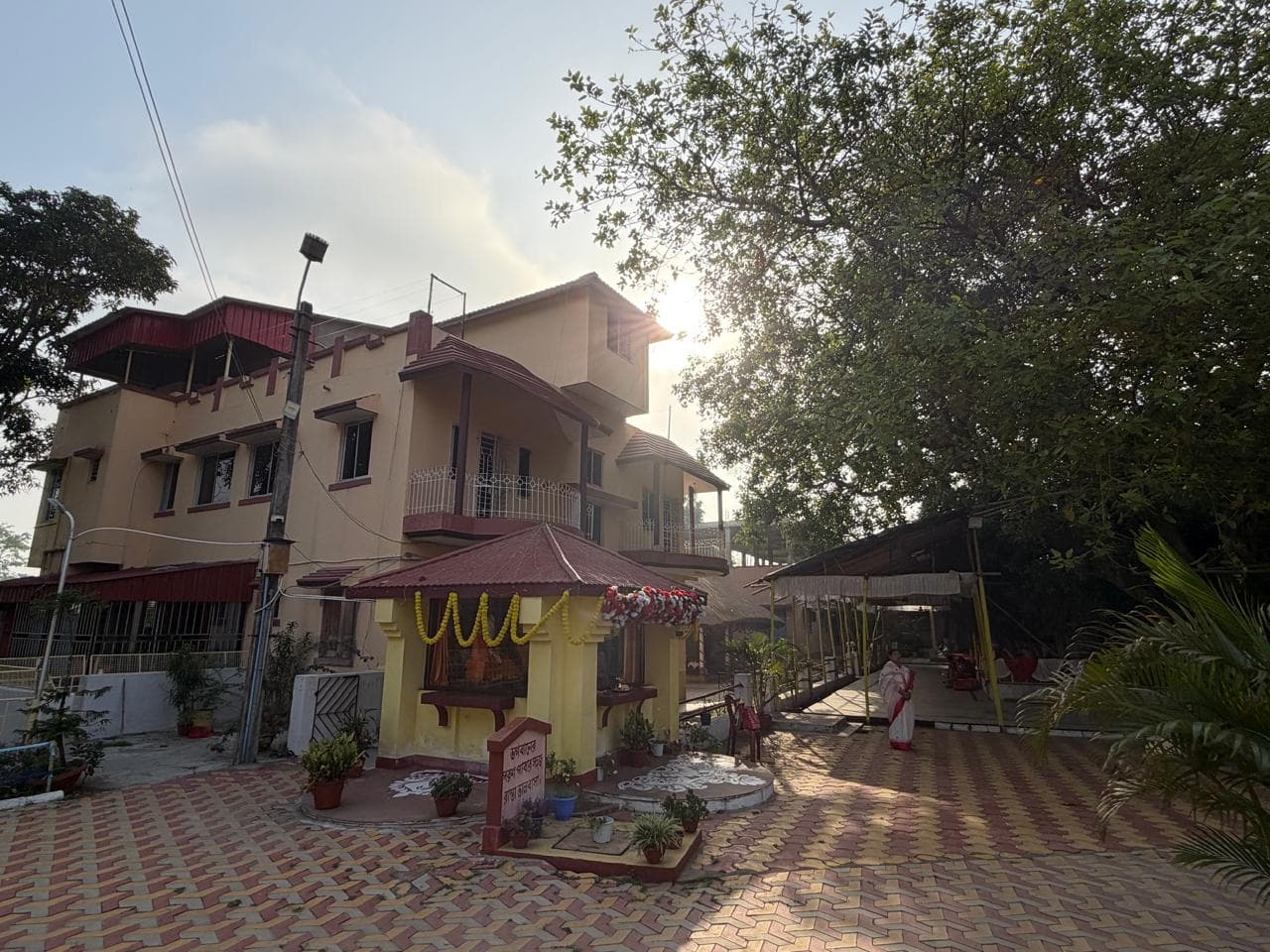 A yellow two-storey building and a small red-roofed shrine on a paved courtyard, with the sun low behind the trees