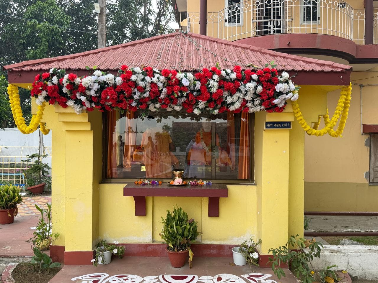 A small yellow shrine with a red roof, crowned with red and white flowers and hung with marigold garlands
