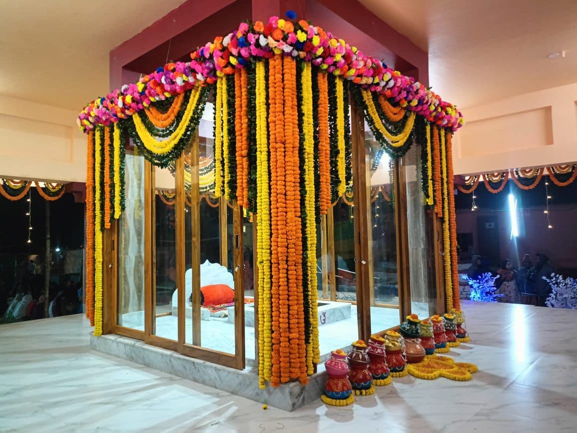 A glass-walled shrine hung with long marigold garlands on a marble floor, with offerings in small pots at its base