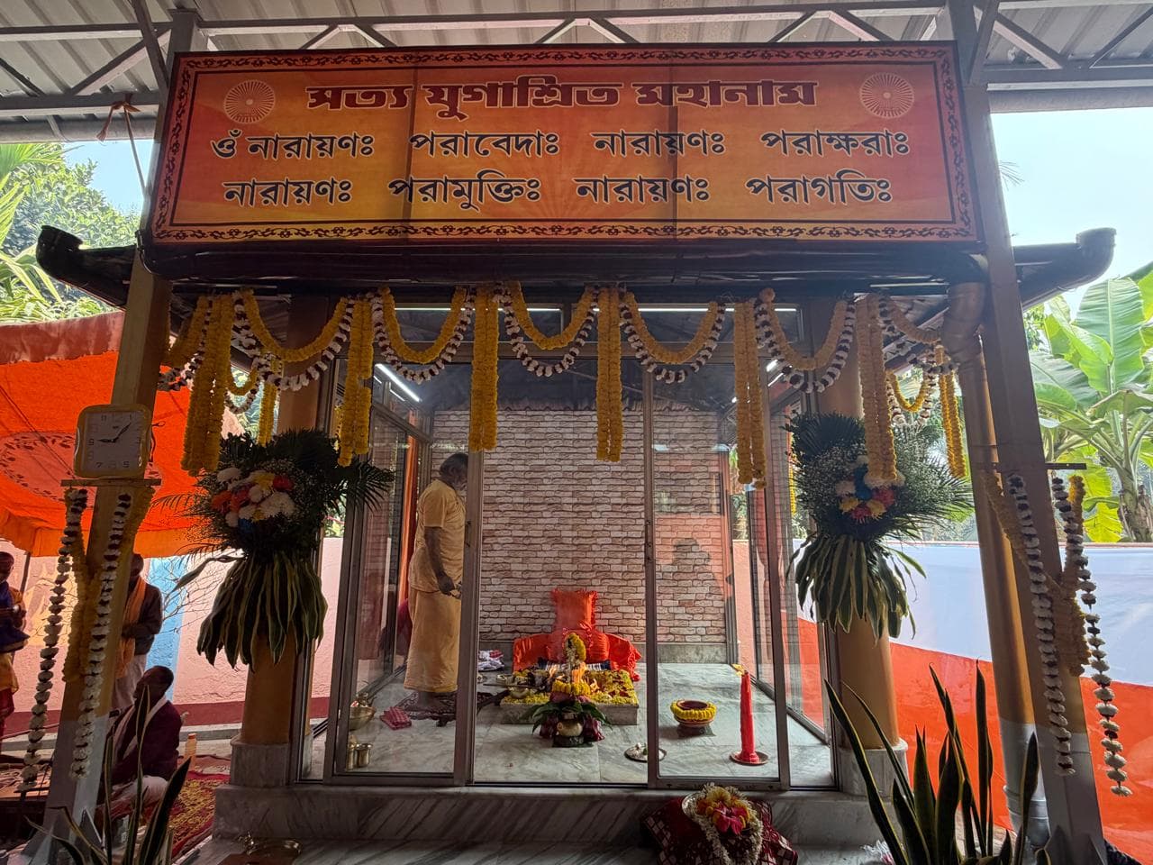 A small glass shrine under a canopy bearing an inscription in Bengali script, garlanded with marigolds