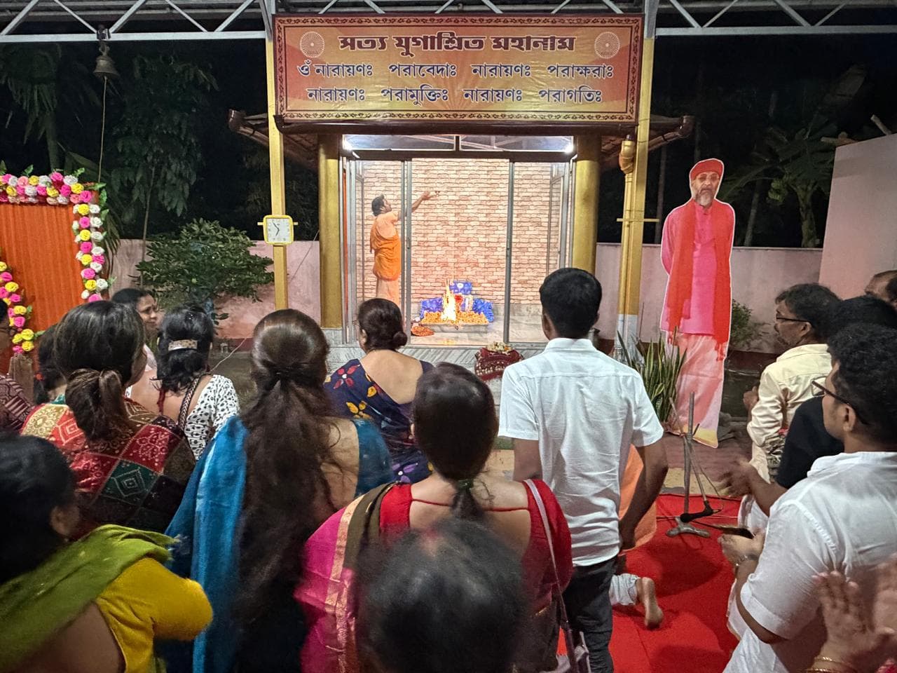 People standing at night before a small garlanded shrine beneath a canopy inscribed in Bengali script