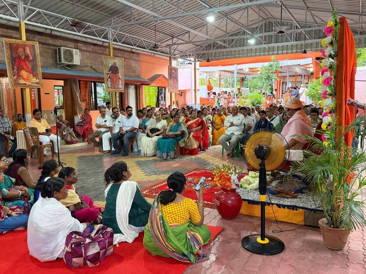 A seated gathering on carpets and chairs under a high metal-roofed hall, facing a decorated dais