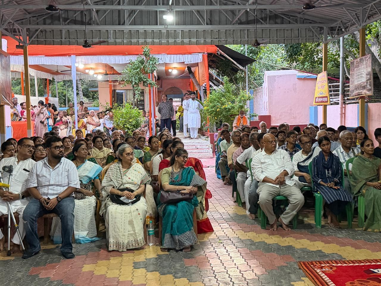 Rows of people seated on chairs in a covered courtyard, with more people standing at the back