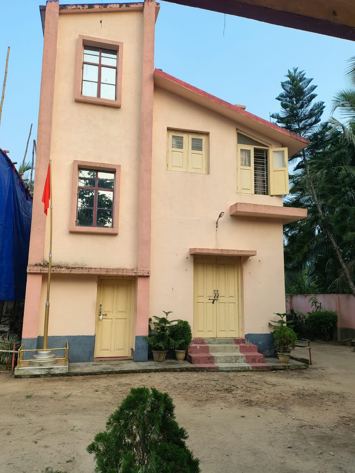 A tall two-storey pink building with yellow doors, a red flag on a pole beside it and a sandy yard in front