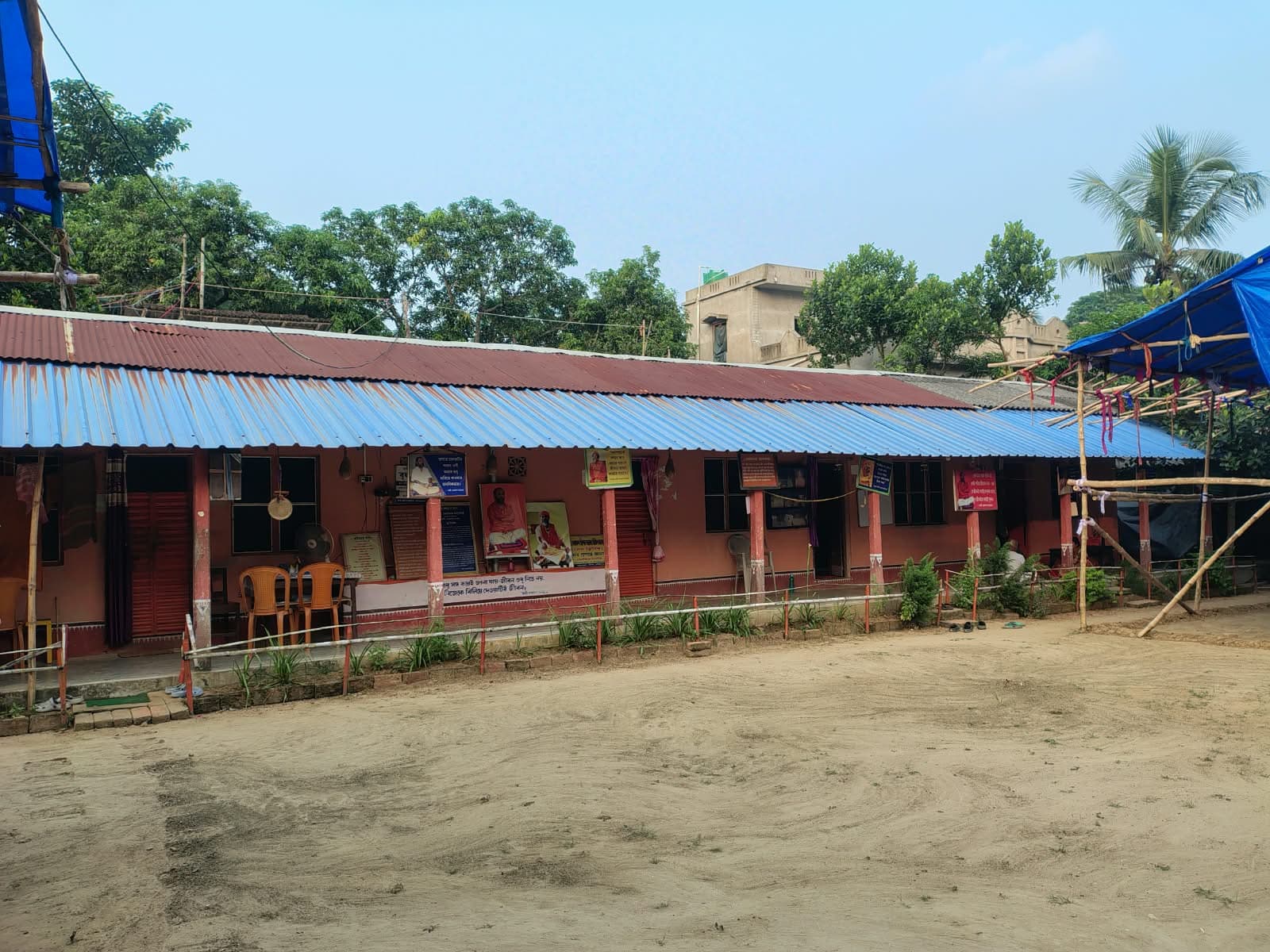 A long single-storey building with a blue tin roof and a covered veranda across an open sandy yard