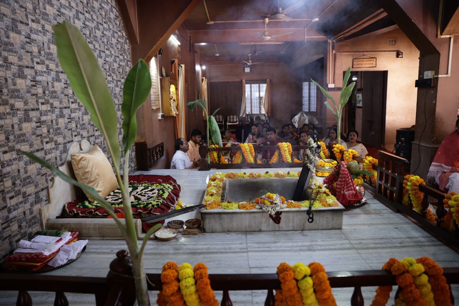 A wide view of a fire altar edged with marigold garlands in a wood-panelled hall, with people seated around it
