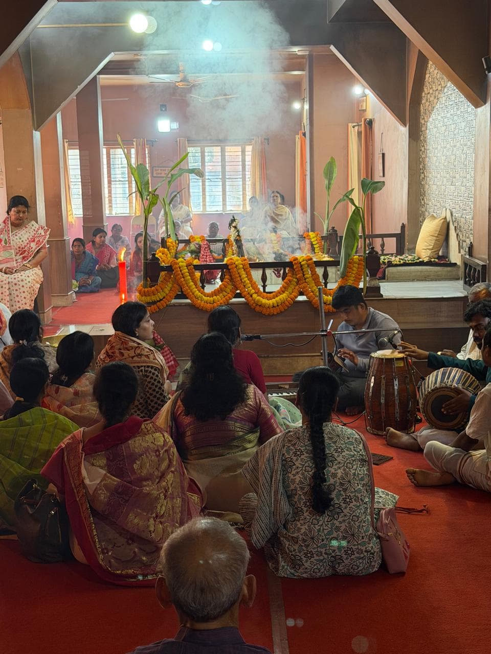 Women seated on a red floor facing a garlanded fire altar, with smoke rising in the hall