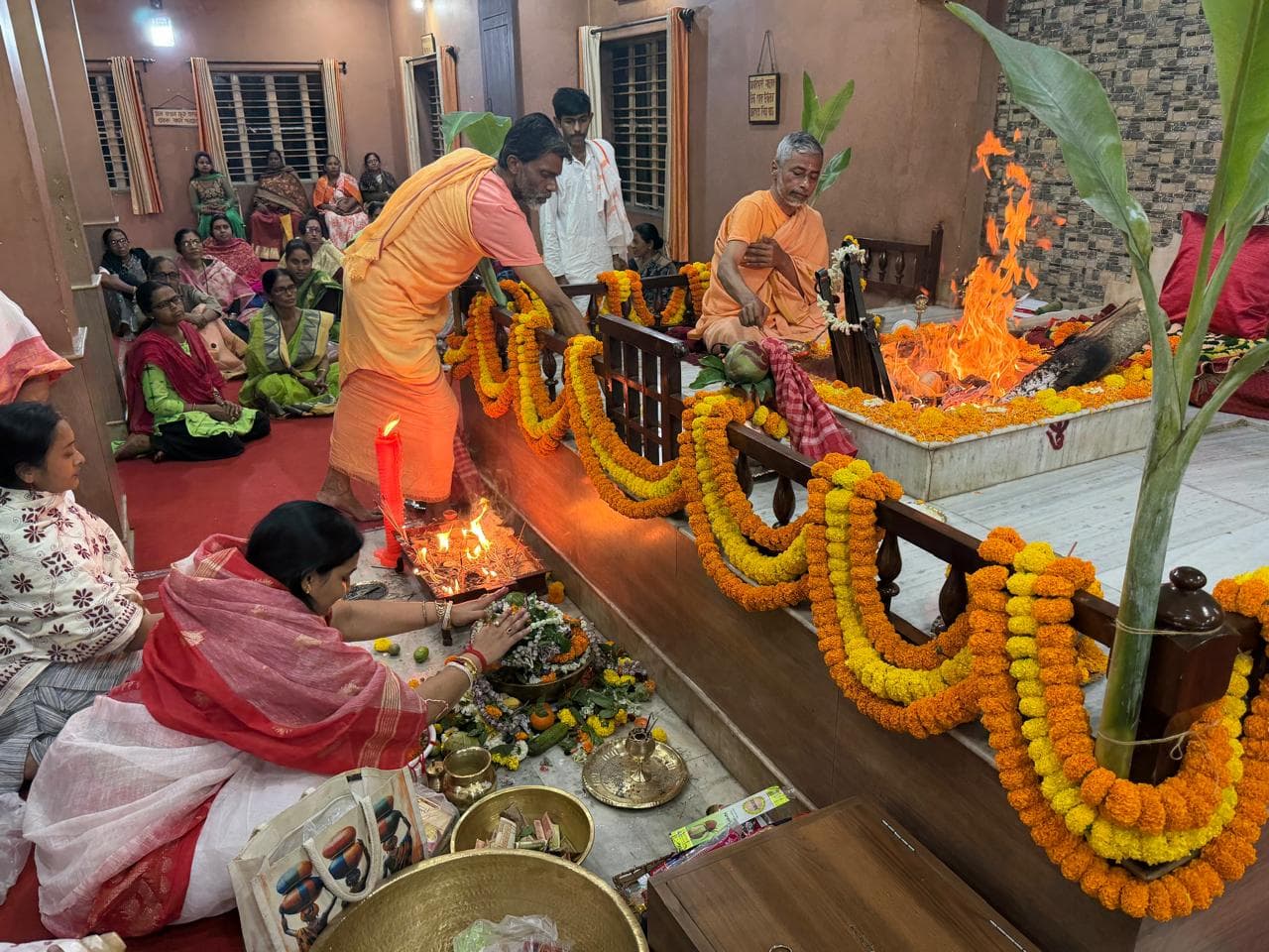 A ritual at a fire altar, with flames rising and people in saffron and white making offerings beside marigold garlands