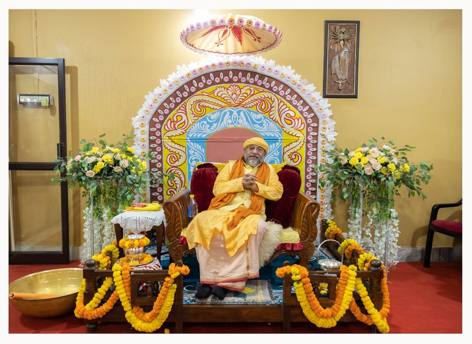 A figure in saffron robes seated on an ornate throne before an arched, painted backdrop, flanked by flower arrangements