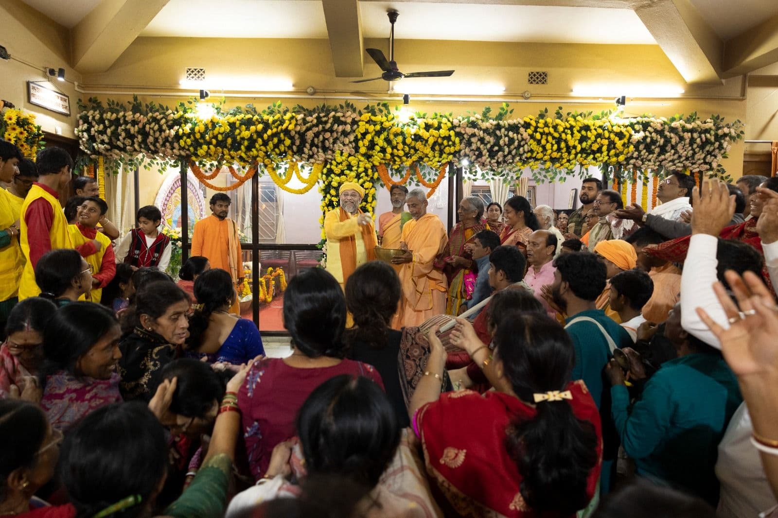 A crowded hall of people with raised hands facing a stage decorated with yellow and white flowers