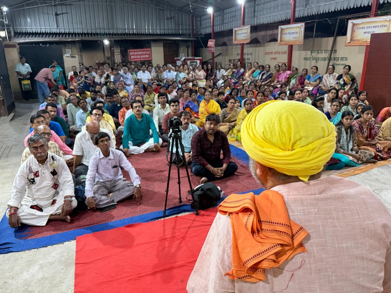 A large seated audience in a hall, seen from behind a person in a yellow turban, with a camera on a tripod