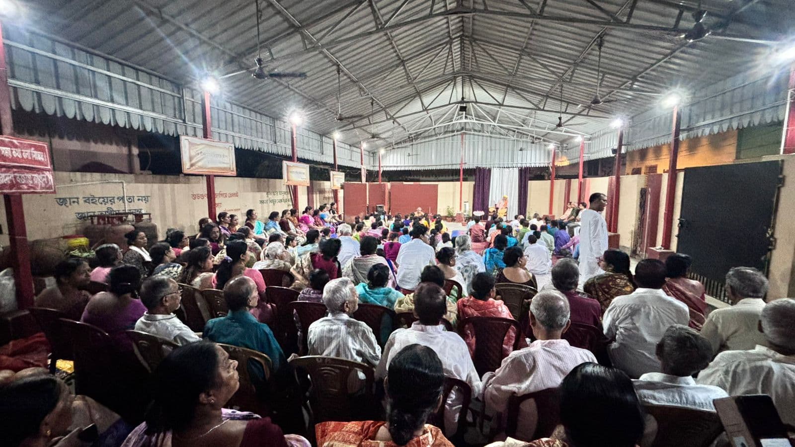 A large audience seated on chairs under a high metal-roofed hall