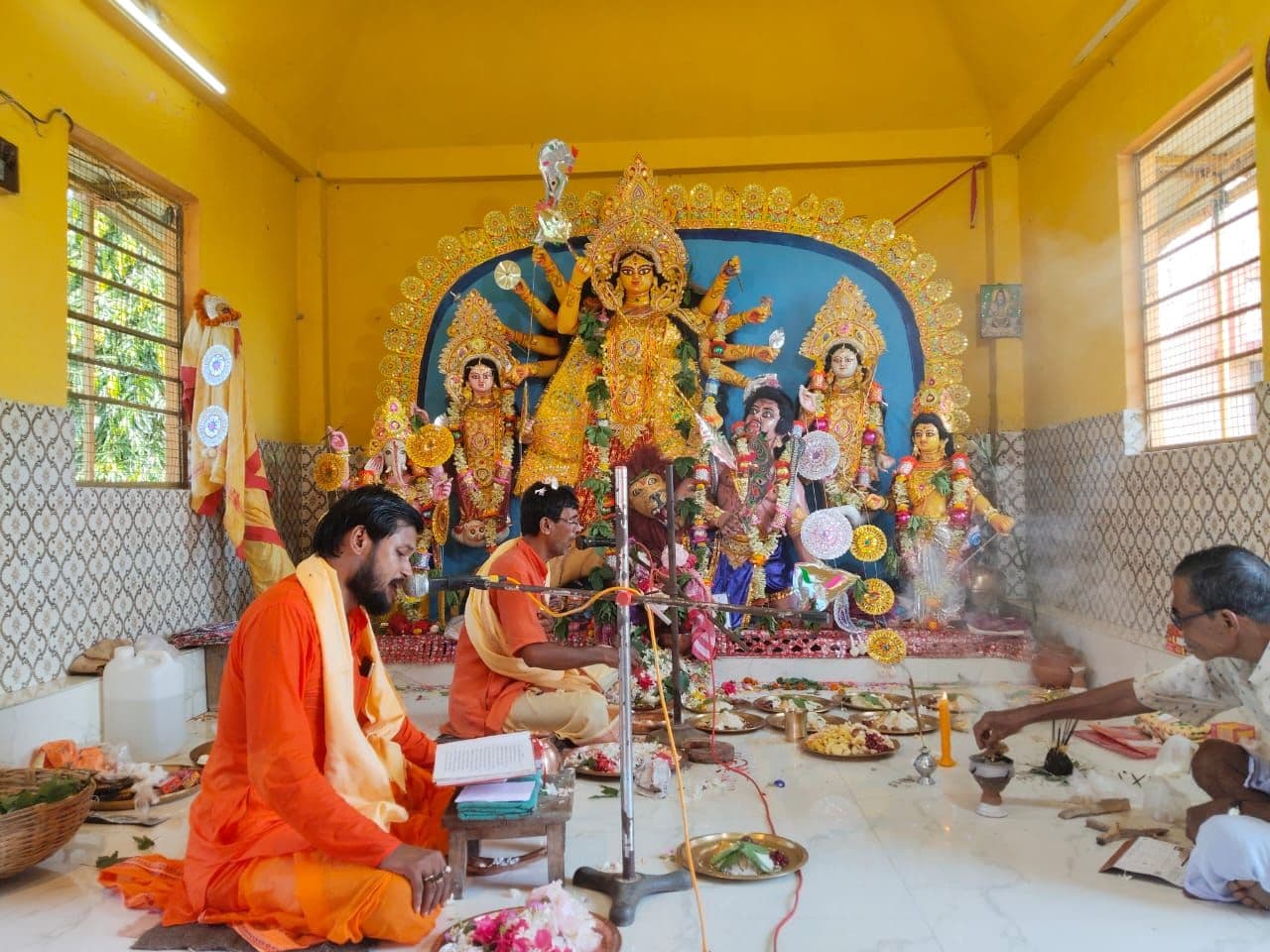 An elaborate idol of a many-armed goddess in a yellow shrine room, with a priest in saffron performing a ritual before it