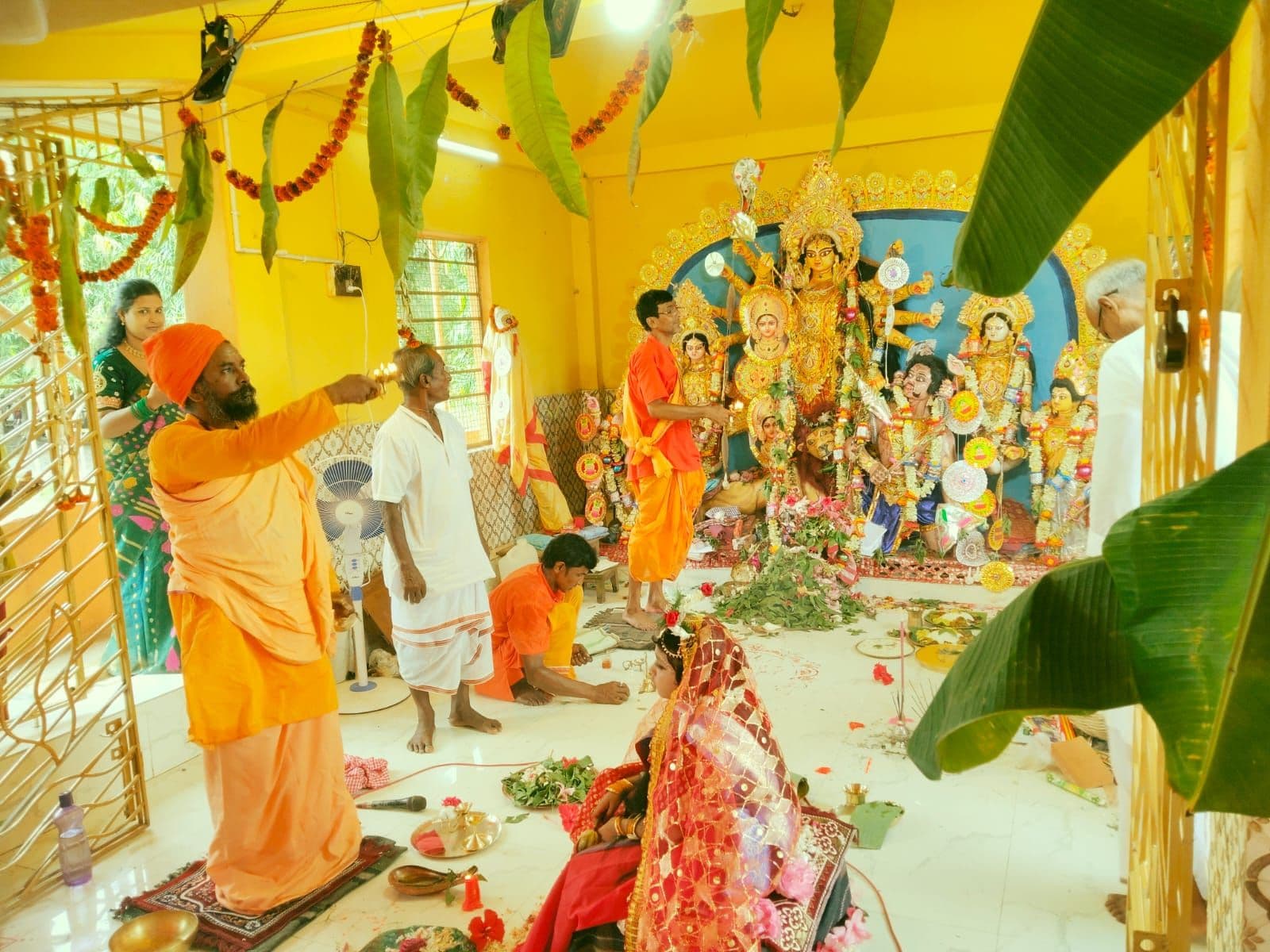 People in saffron and white performing a ritual before a many-armed goddess idol in a yellow shrine room