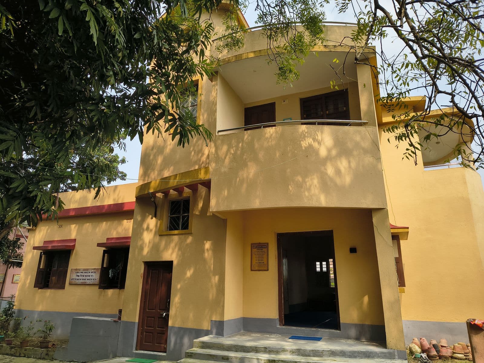 A two-storey yellow building with a balcony, partly shaded by trees