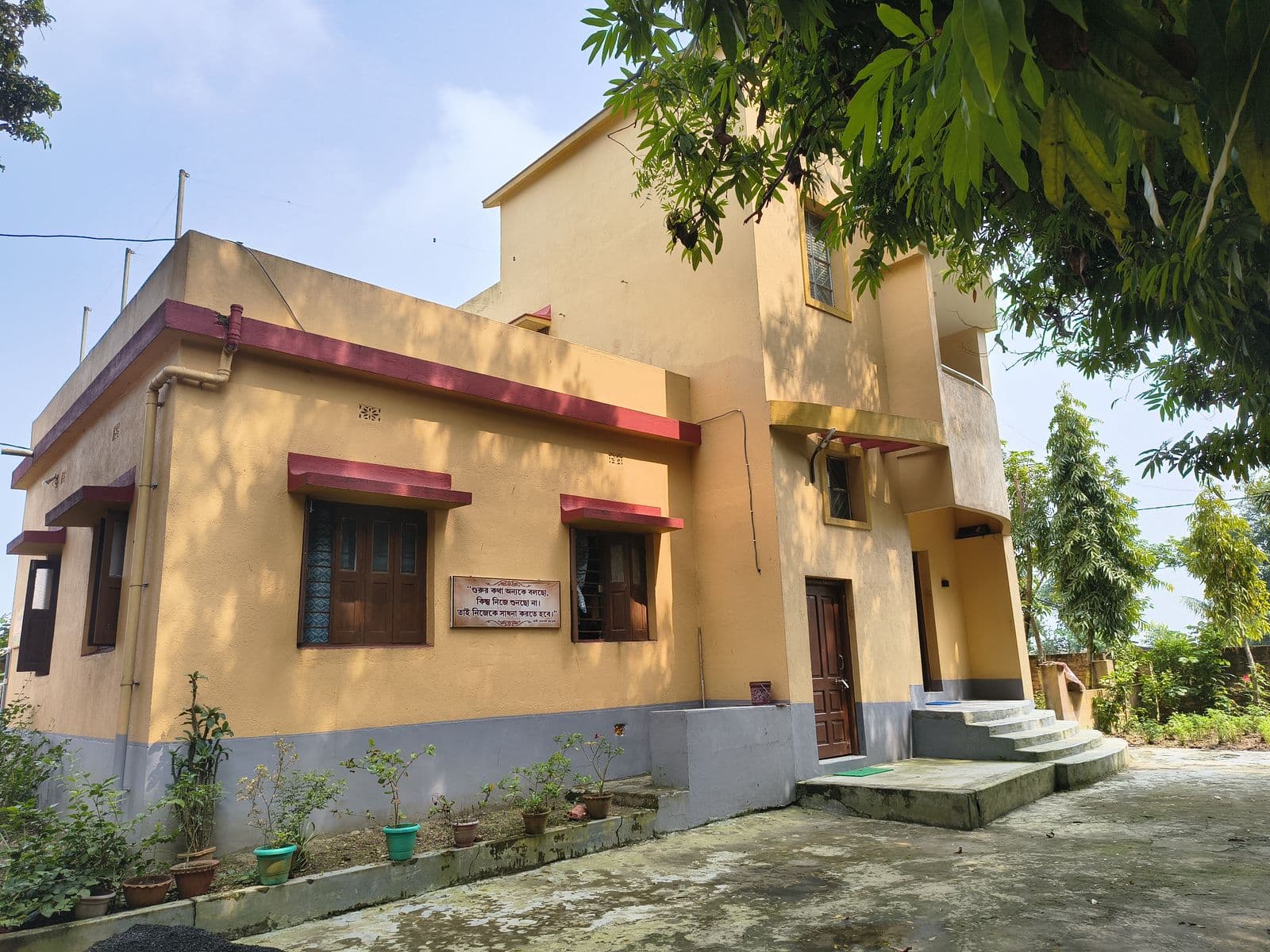 A single-storey yellow building with red window ledges and steps to its door, under shady trees