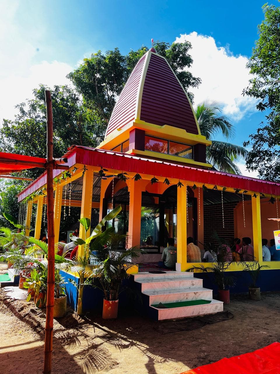 A temple with a pointed maroon spire and a red-and-yellow roof over an open hall, among palm trees