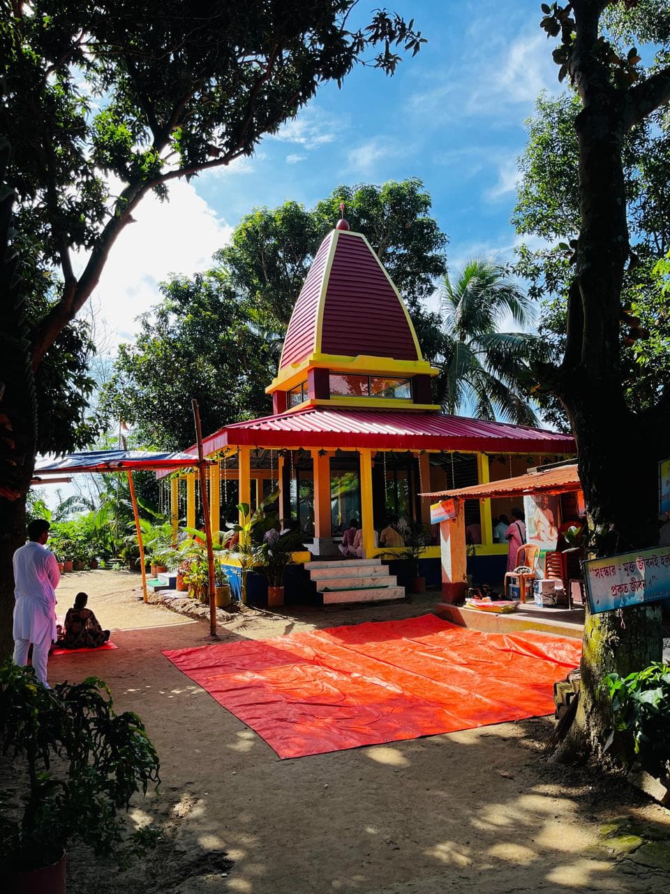 A temple with a maroon spire beyond a red carpet laid on a sandy path, framed by tall trees