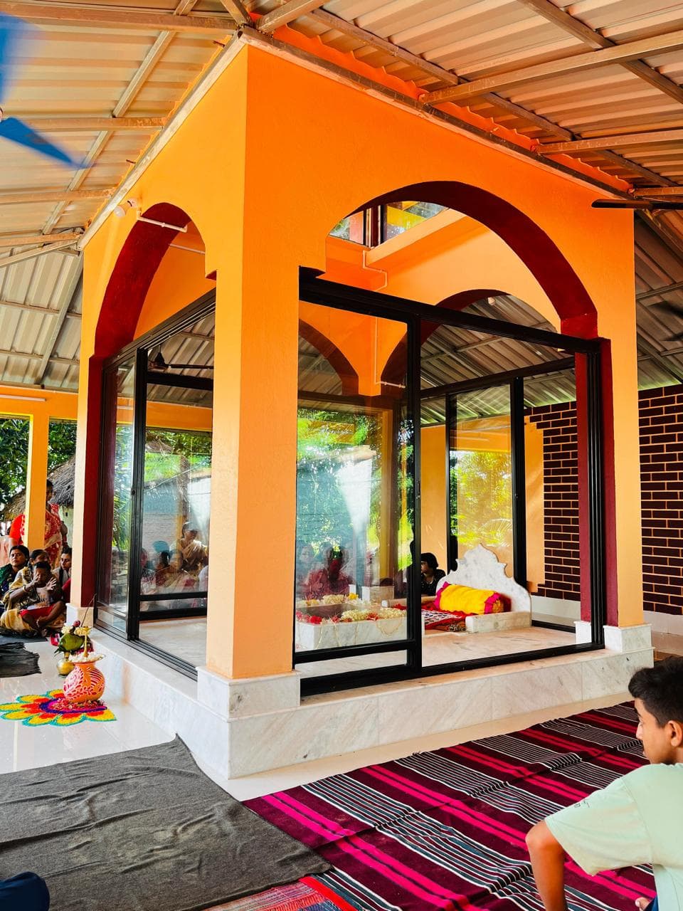 A glass shrine framed by an orange arch inside an open hall, with striped mats on the floor