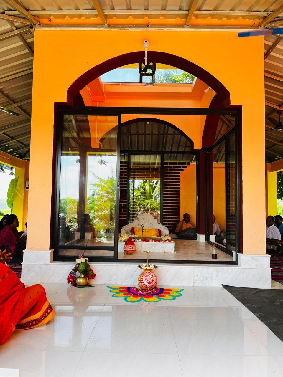 An orange arched shrine with glass walls, with offerings and an alpana design on the white floor in front