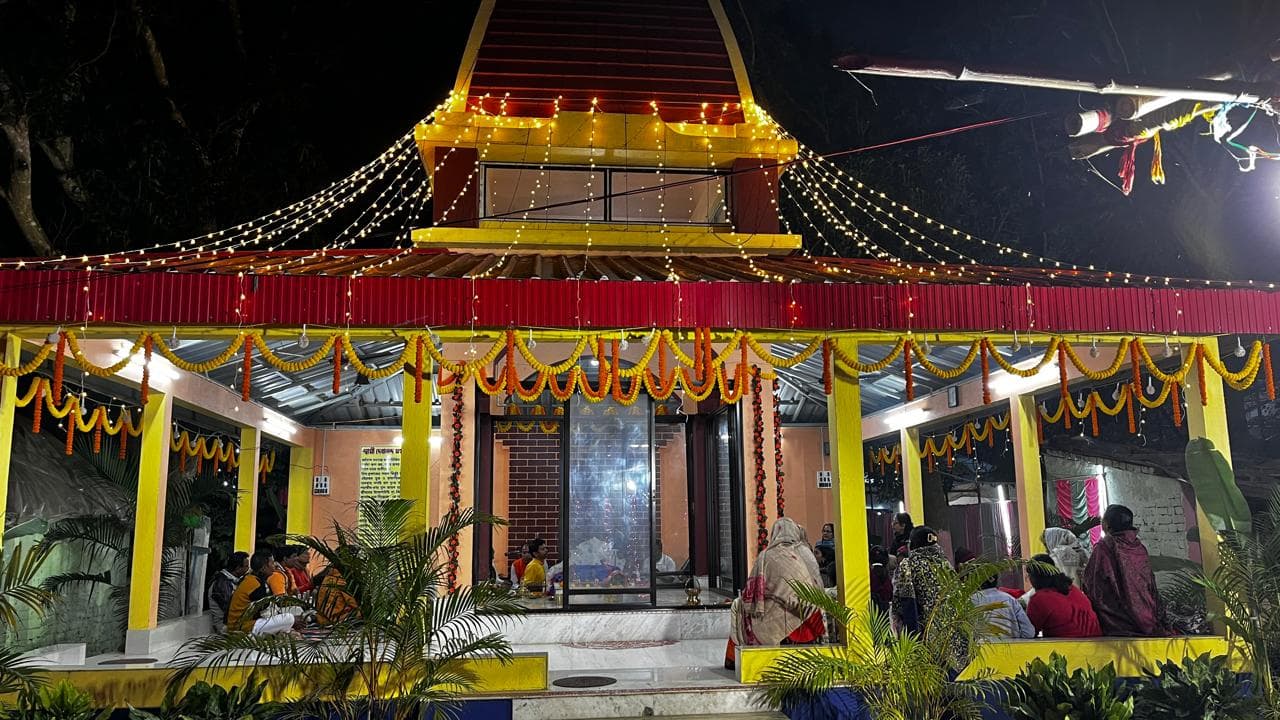 A temple lit with strings of lights at night, its open hall hung with marigold garlands and people seated inside
