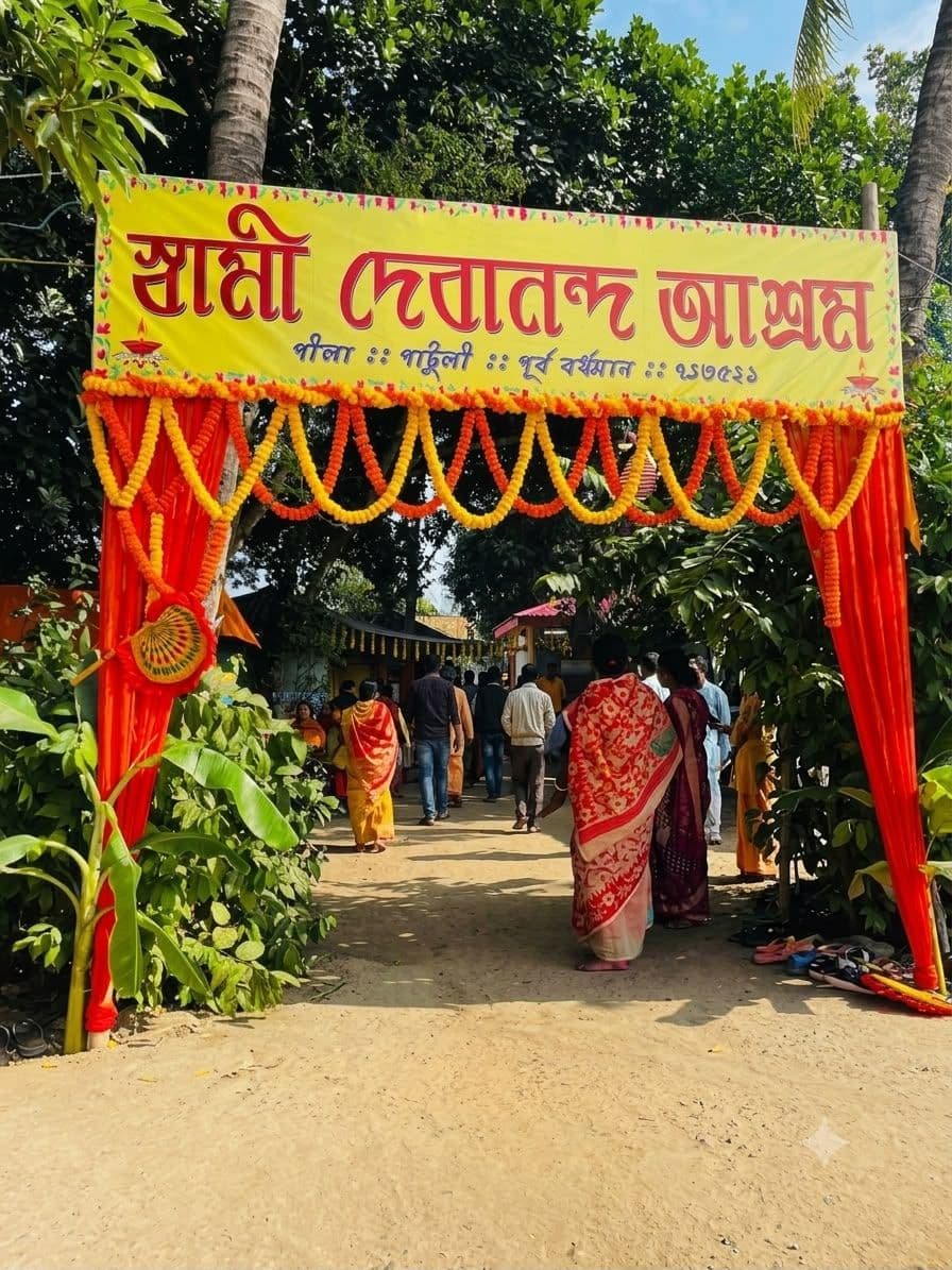 A gateway draped in red and orange cloth and marigold garlands under a yellow banner lettered in Bengali, with people walking through