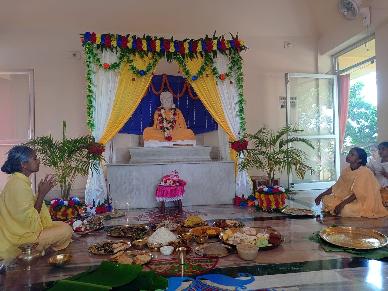 A garlanded altar with a seated statue under draped yellow and white curtains, with offerings of food laid on the floor and two women seated nearby