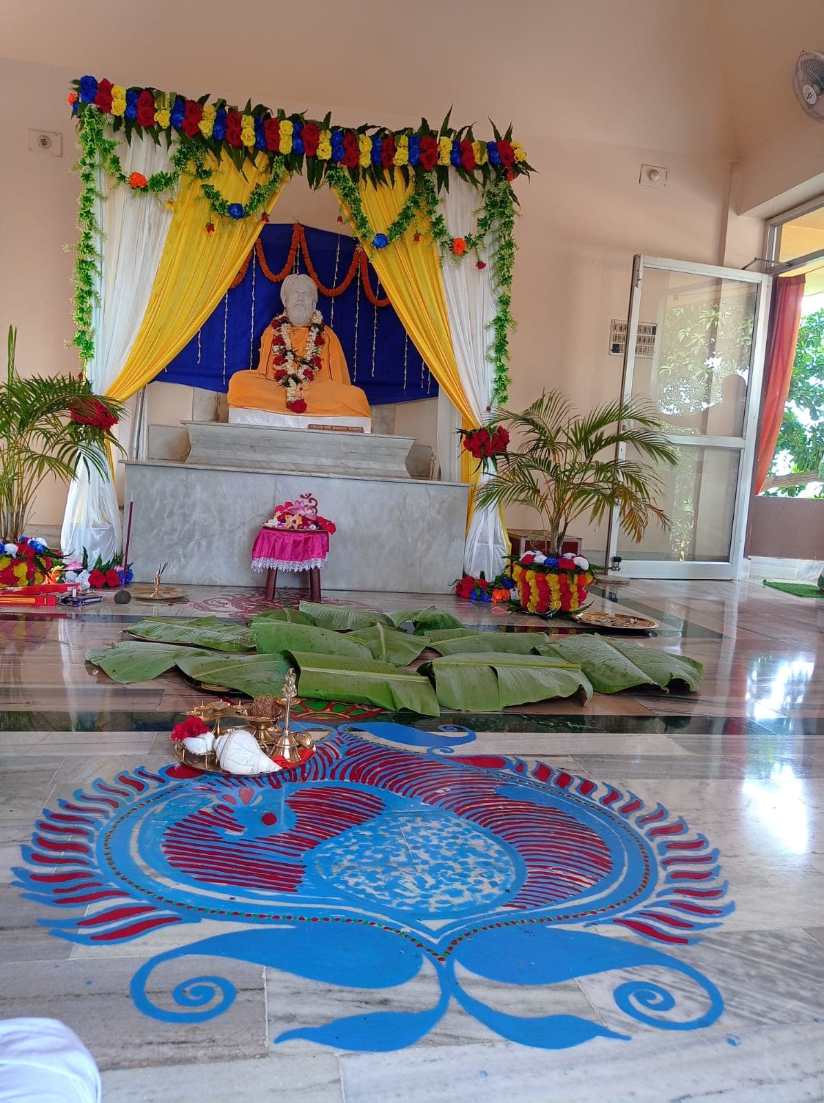 A seated statue on an altar framed by flower garlands and draped curtains, with a large blue alpana design on the floor in front