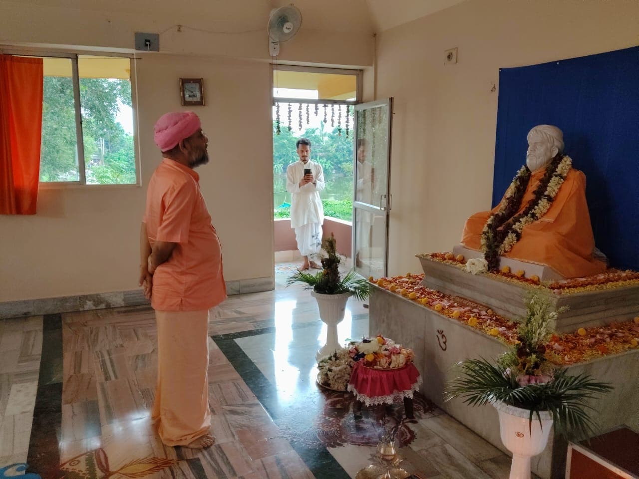 A man in a pink turban standing before a garlanded seated statue against a blue backdrop in a bright hall