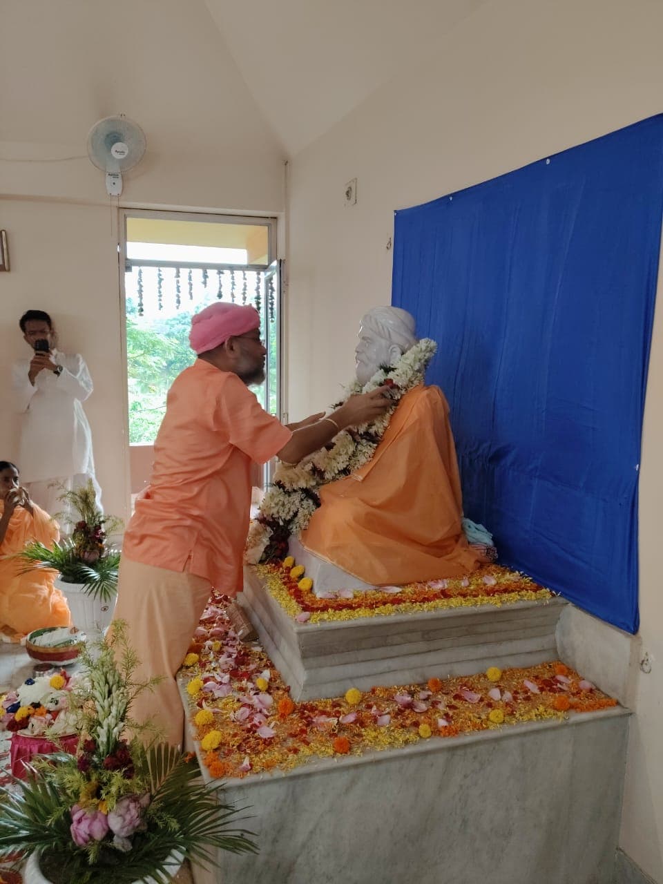 A man in a pink turban placing a garland on a seated statue strewn with marigold petals