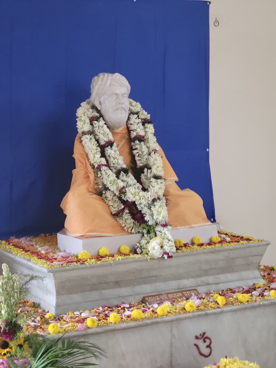 A white seated statue in saffron robes wearing thick flower garlands, on a marble platform scattered with petals