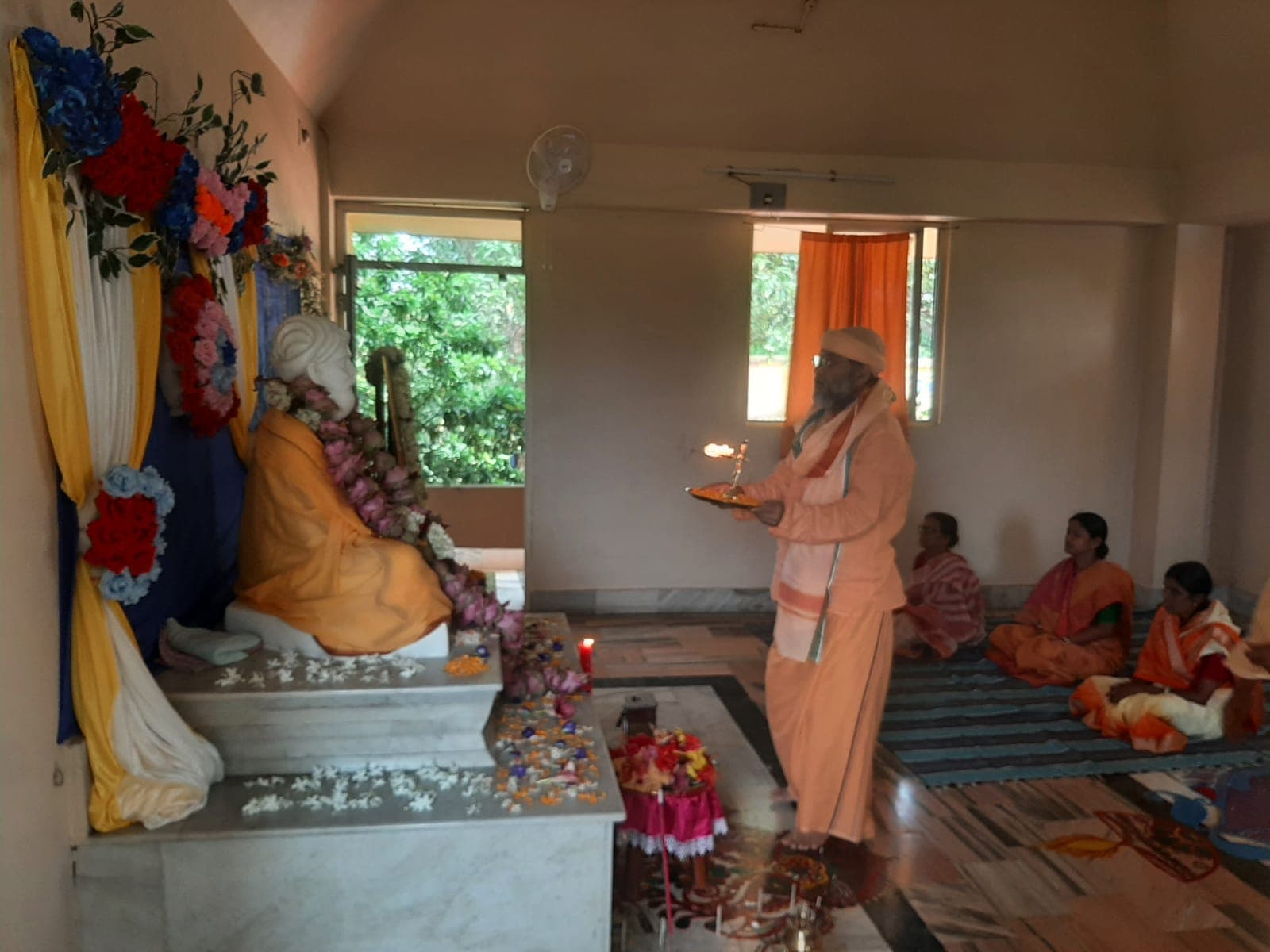 A person in saffron offering a lamp before a garlanded seated statue, with women seated on mats nearby