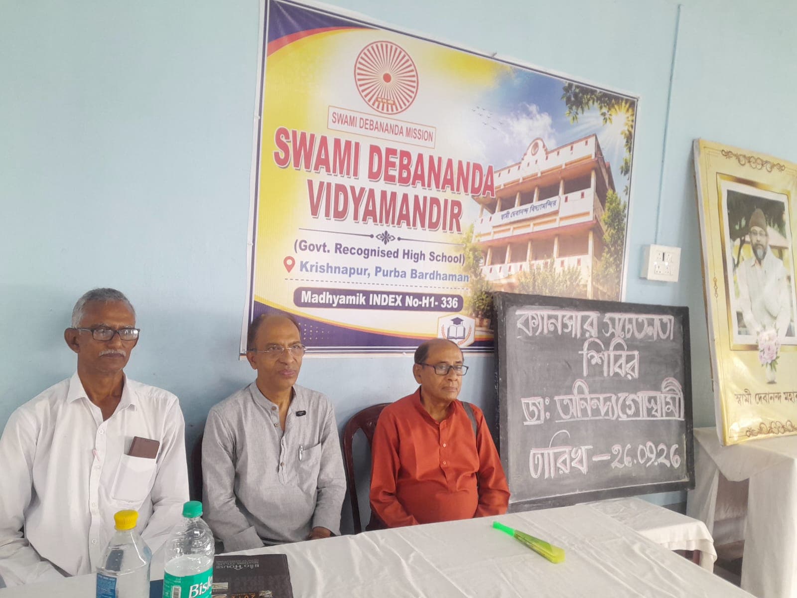 Three men seated at a table in front of a banner of Swami Debananda Vidyamandir and a chalkboard notice in Bengali