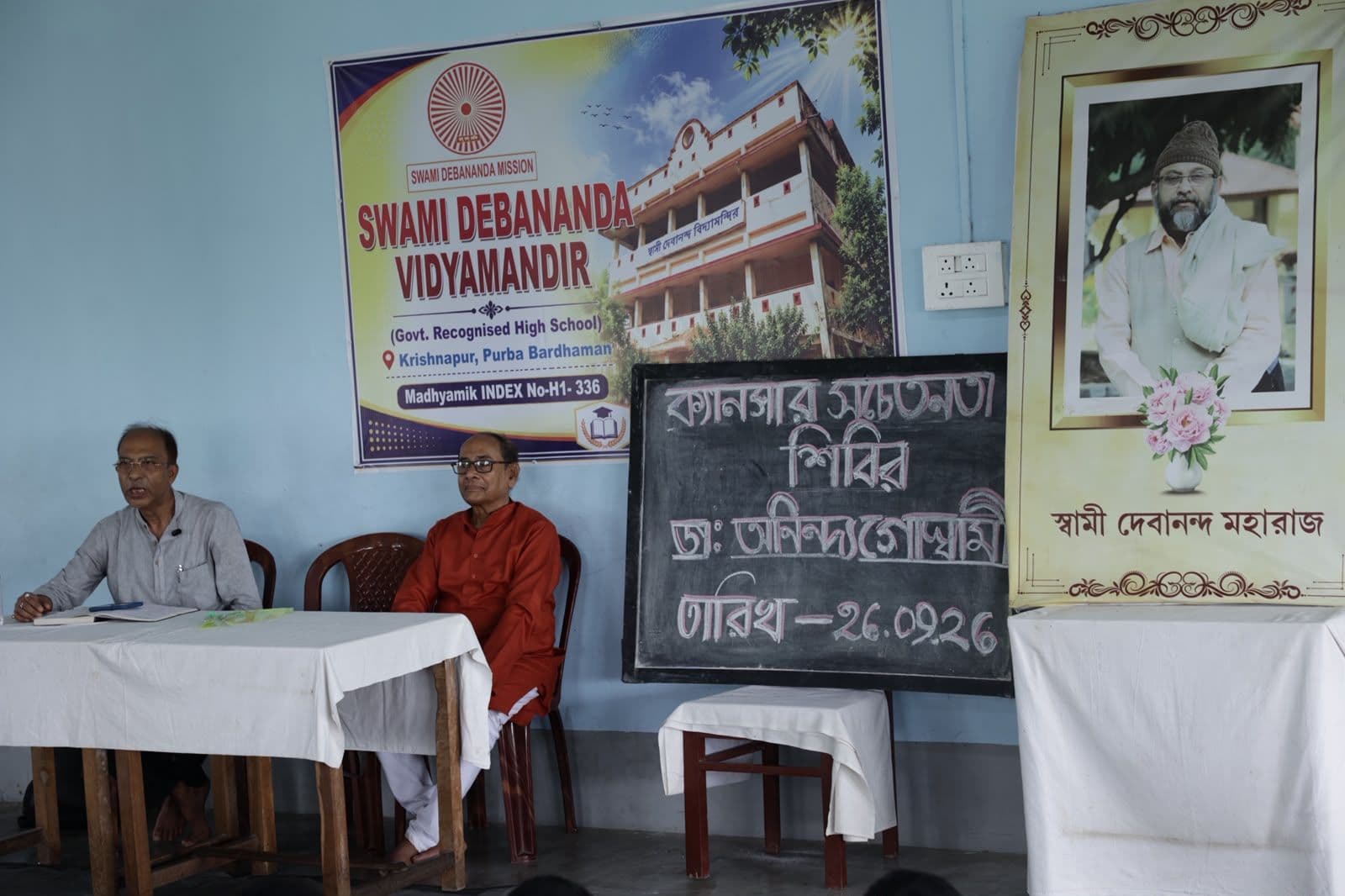 Two men seated at a table beside a chalkboard notice in Bengali and a framed portrait