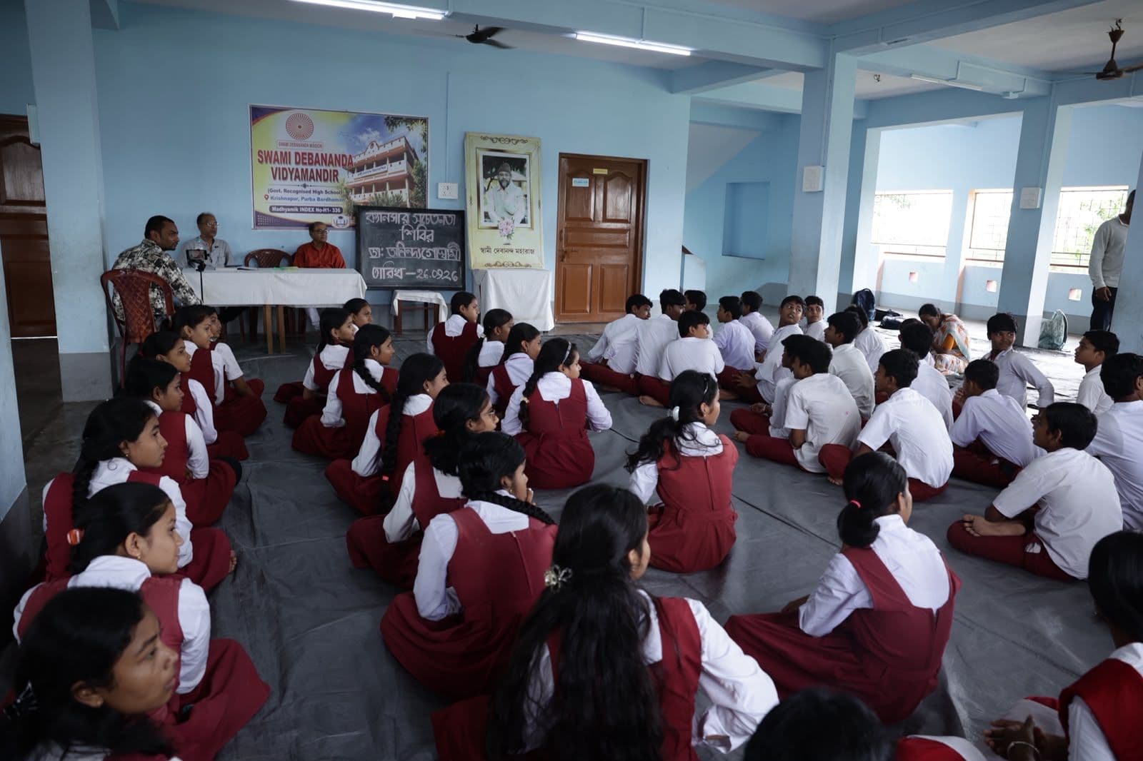 Students in uniform seated on the floor of a hall facing a table at the front